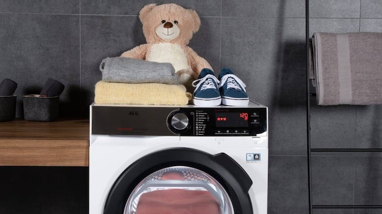 Teddybeer samen met kleding en schoenen boven op een AEG-wasmachine geplaatst.
