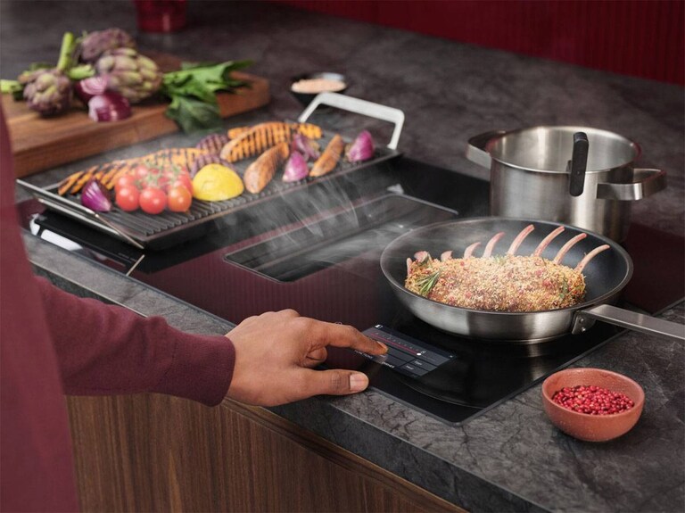 La main d'une personne se dirigeant vers les commandes d'une plaque de cuisson à induction AEG. Sur la plaque de cuisson se trouvent une poêle et une plancha avec des aliments.